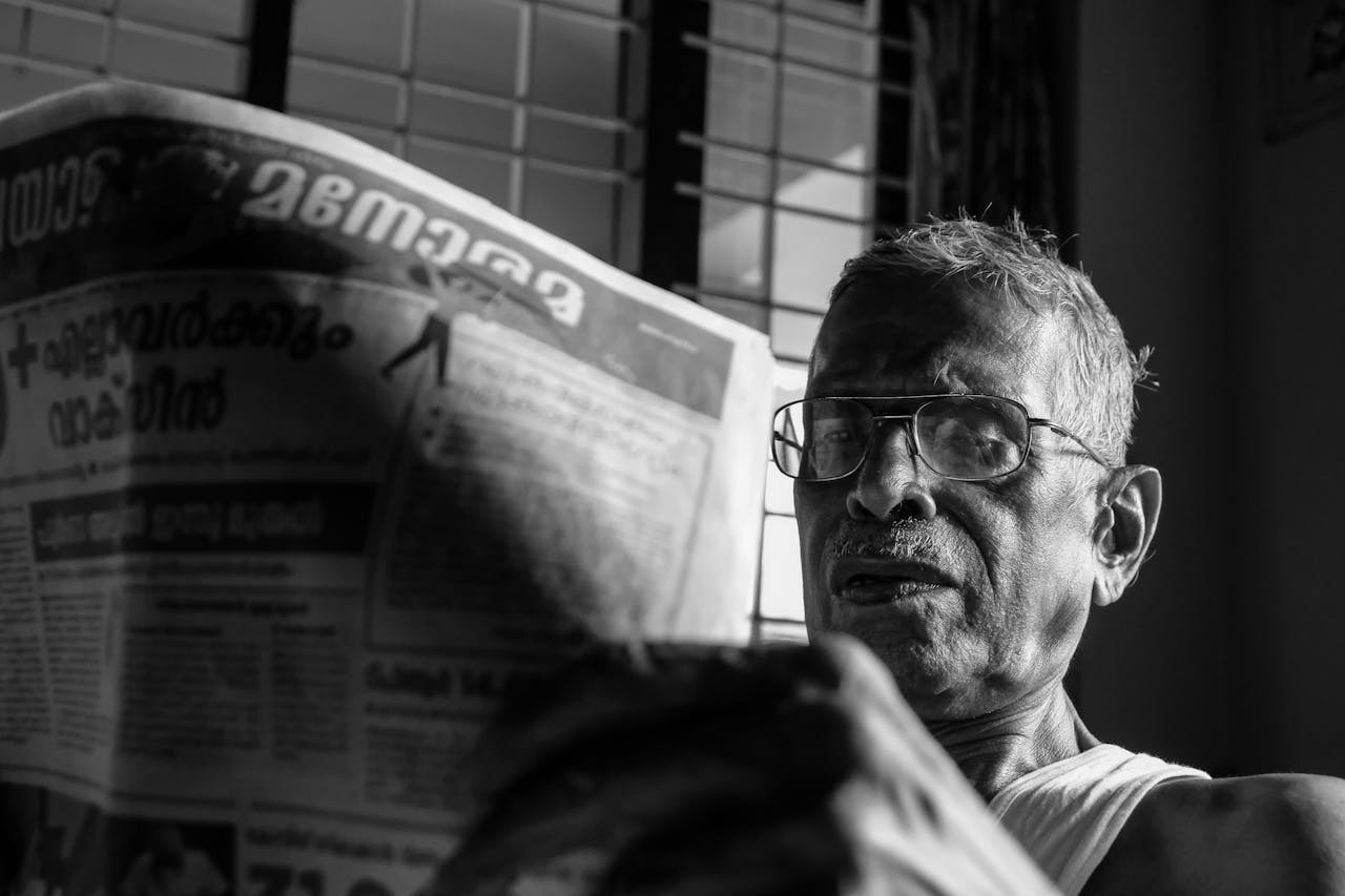 services-img Black and white photo of an elderly man with glasses reading a newspaper in Nilambur, India.